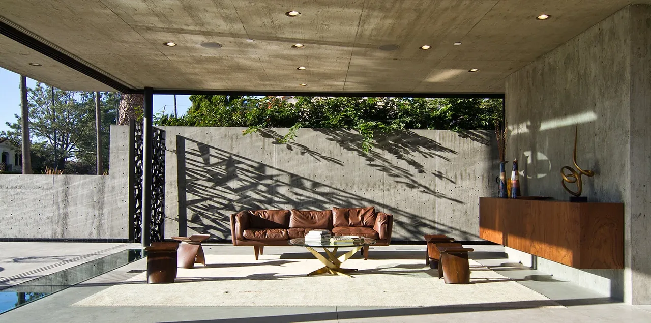 The Cresta living area features concrete walls and ceiling, a leather sofa, glass walls, and a sculptural table base.