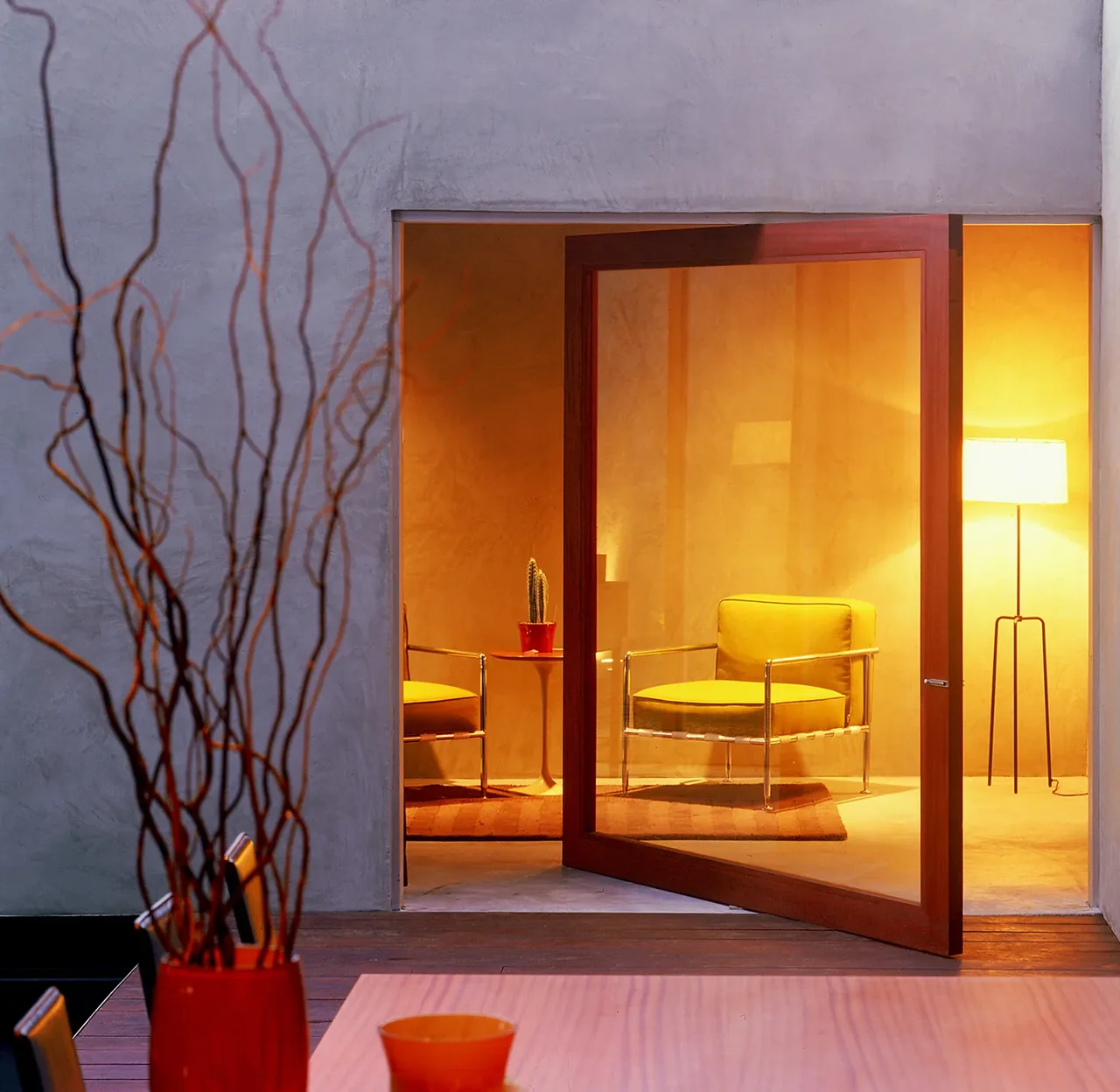 Interior view through an open pivot door showing a modern seating area with yellow chairs and a floor lamp.