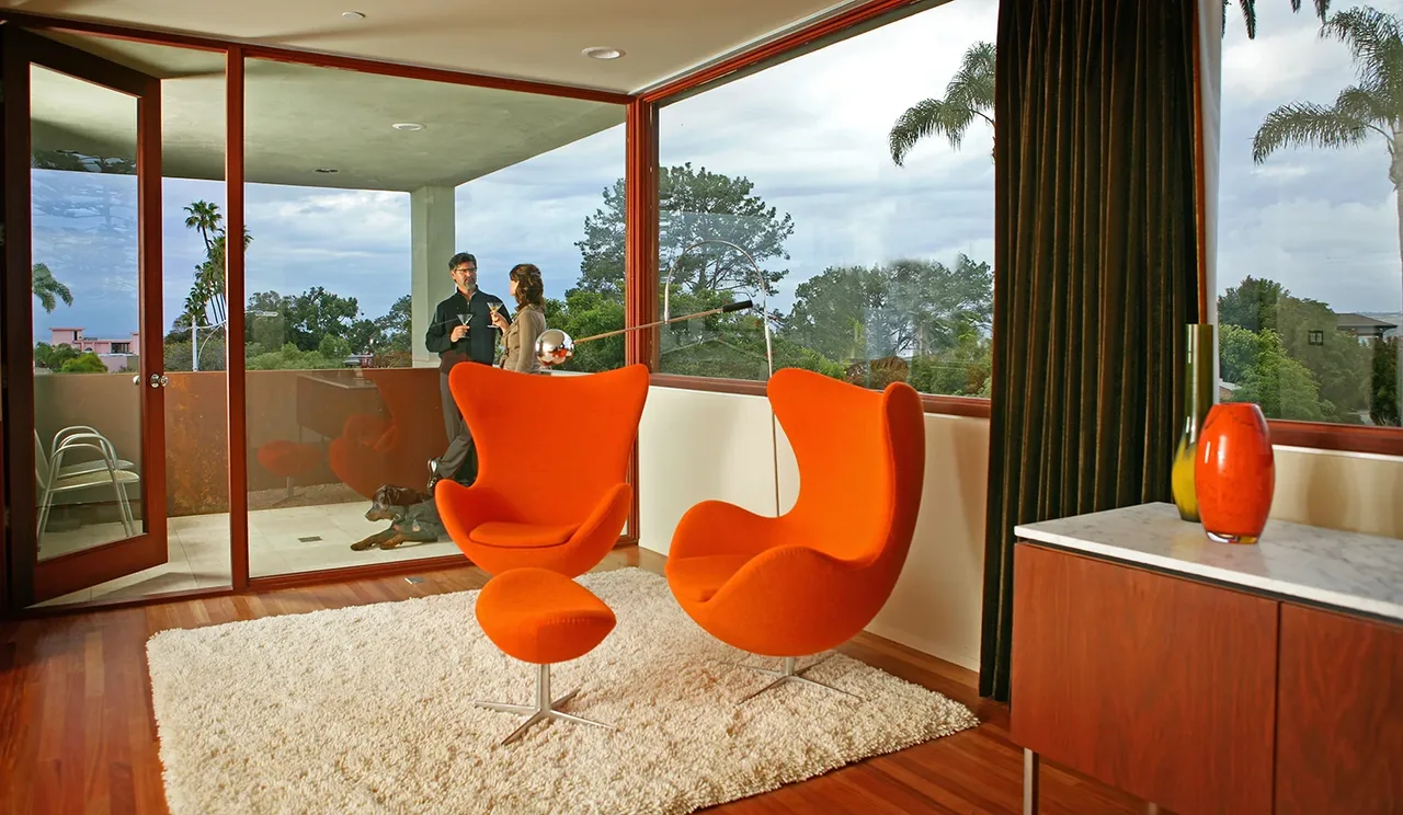 Modern living room corner with orange armchairs, hardwood flooring, and floor-to-ceiling glazing opening to an exterior deck.