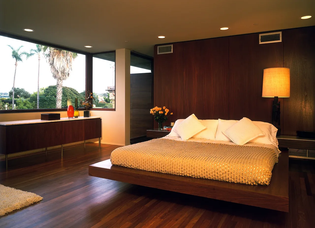 Bedroom featuring a modern wood-paneled wall, large corner windows, and a low platform bed with white bedding.