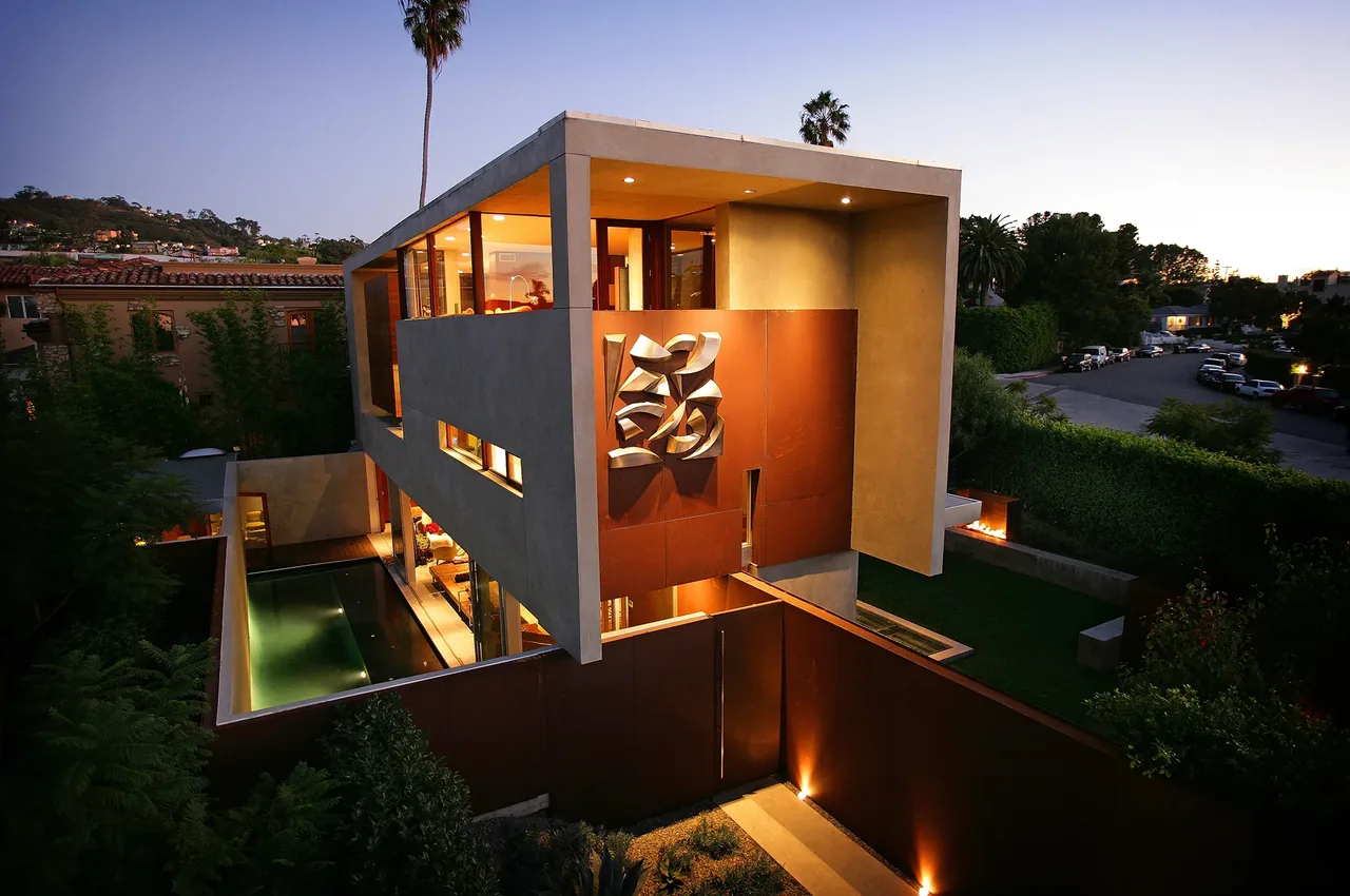 Modern two-story house at dusk with a reflecting pool and illuminated metal wall art on the exterior.