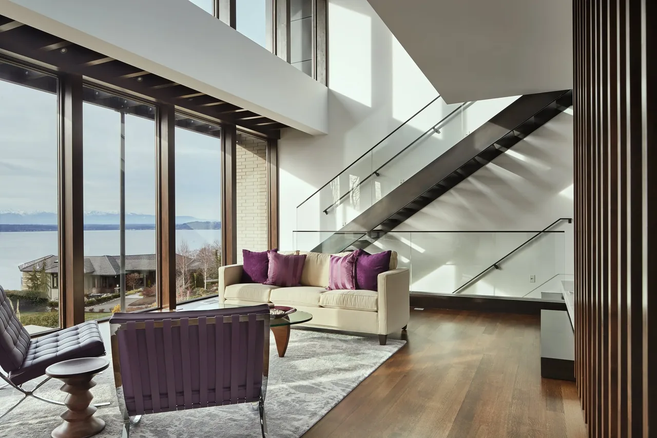Living room with floor-to-ceiling windows reveals a view of water and mountains, featuring a floating metal staircase and purple-accented seating.