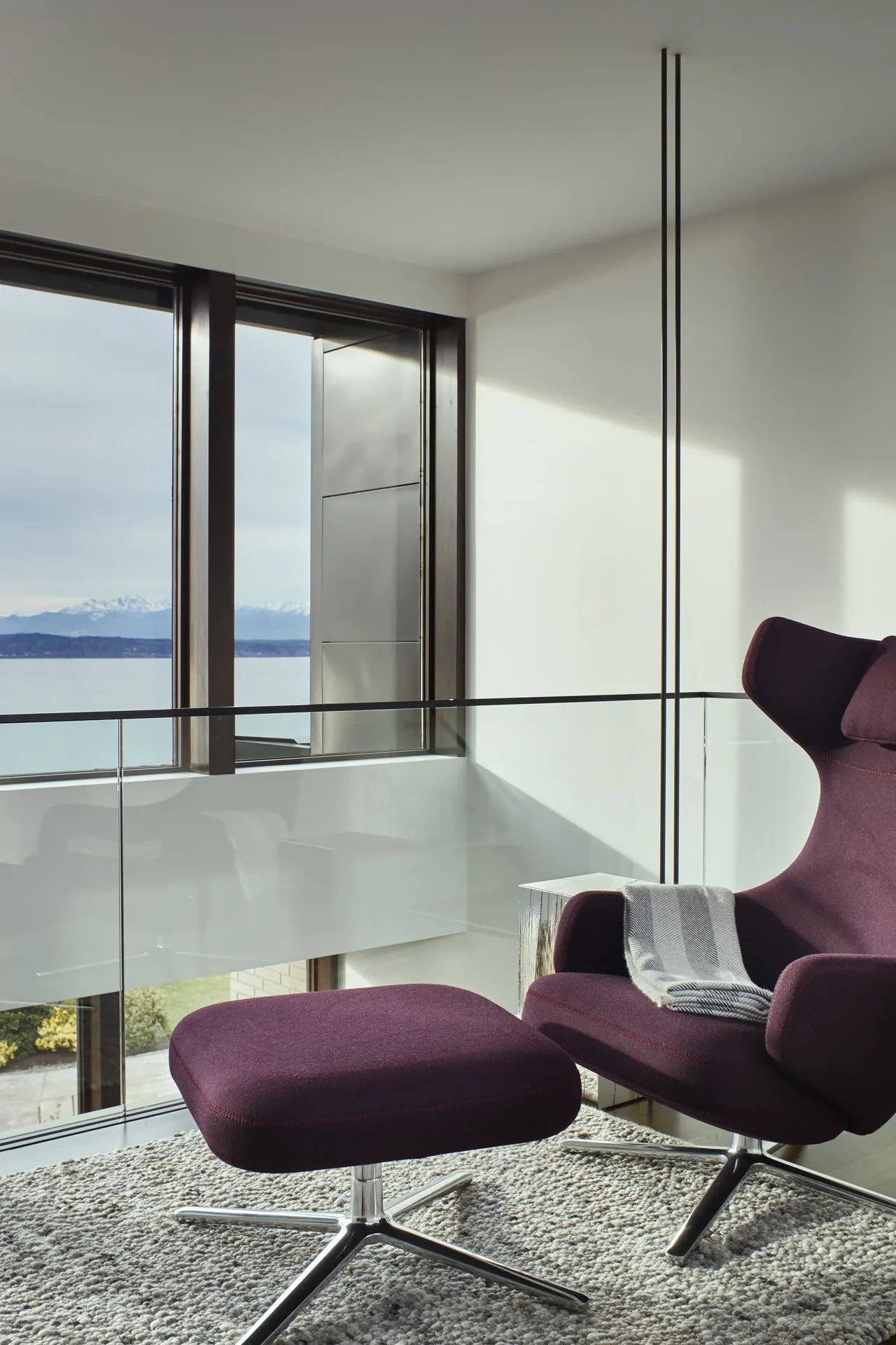 Seating area with a purple lounge chair and ottoman beside a glass railing offering a water view with mountains in the distance.
