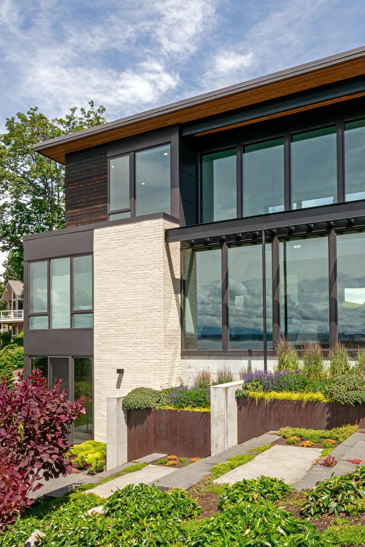 The Woodway house features large glass walls, vertical wood paneling, and rust-colored metal planters integrated into the landscape.