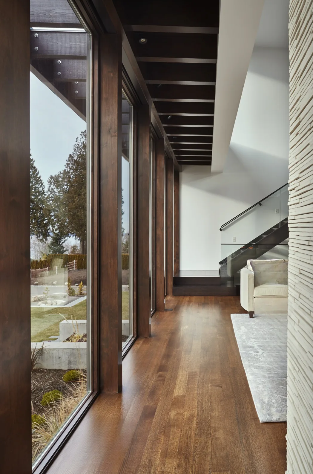 Interior hallway with floor-to-ceiling glazing and dark wood floors adjacent to a living area with a textured wall and glass-railing staircase.