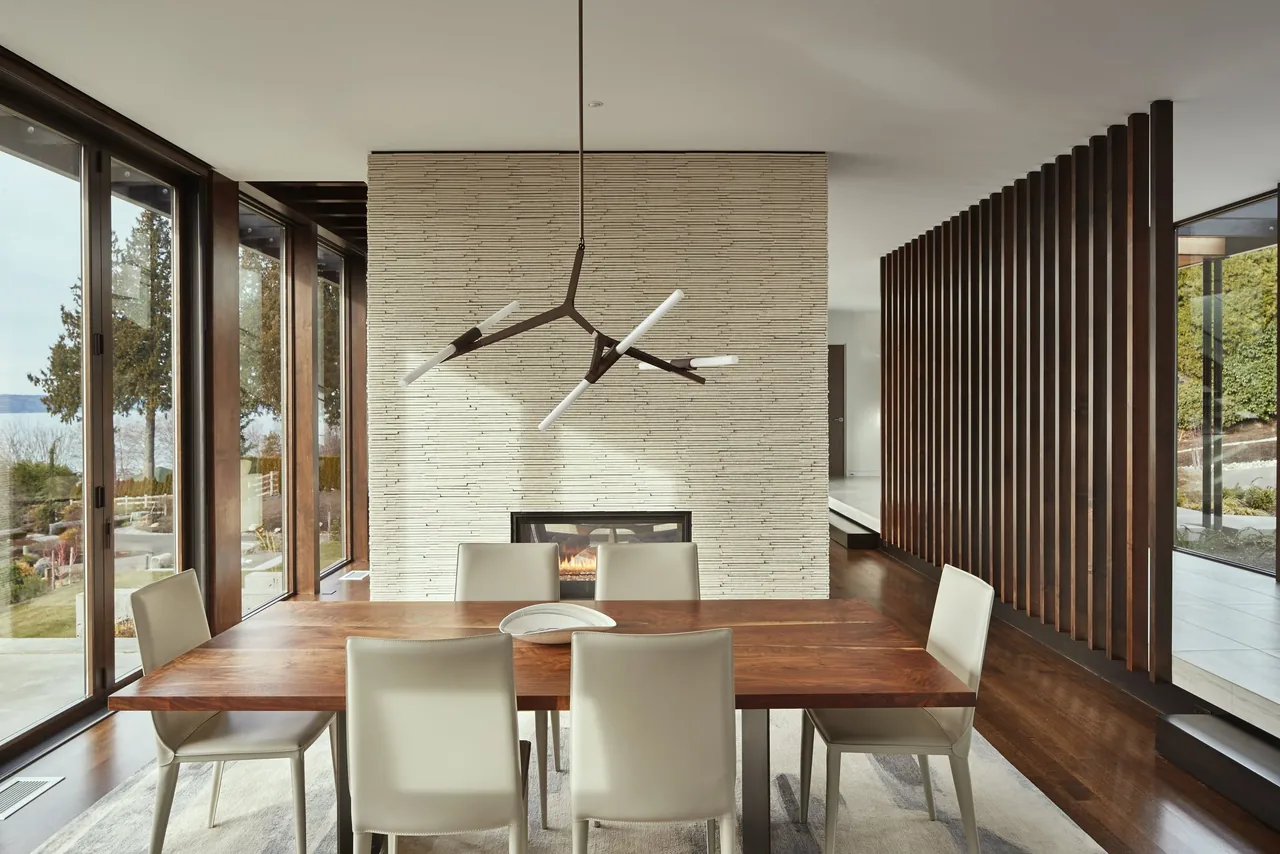Modern dining area with a wood table, cream leather chairs, and a linear fireplace centered on a textured white wall with large glass window walls.