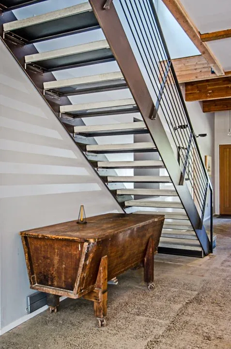 The hallway features a modern metal open-riser staircase with a cable railing and a rustic wooden chest on small wheels beneath it.
