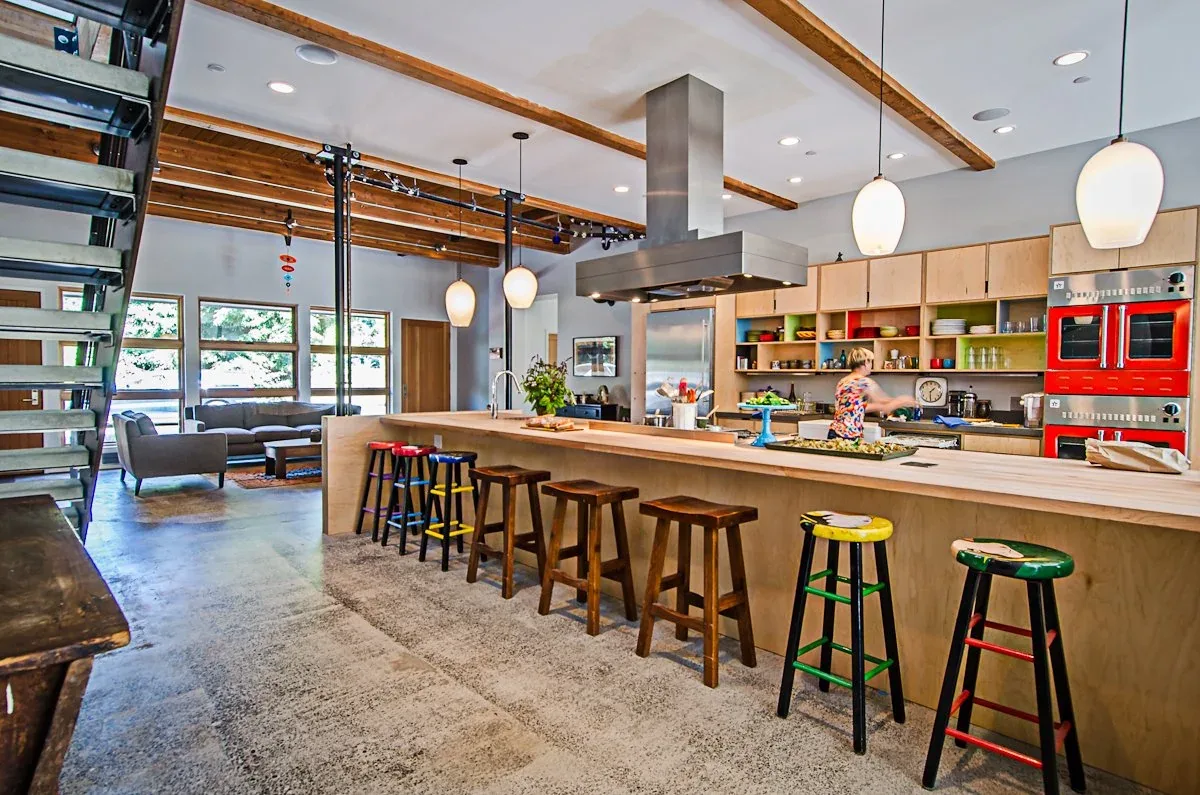 Open kitchen with exposed wooden beams, colorful stools, and a metal range hood adjacent to a living area with large windows.