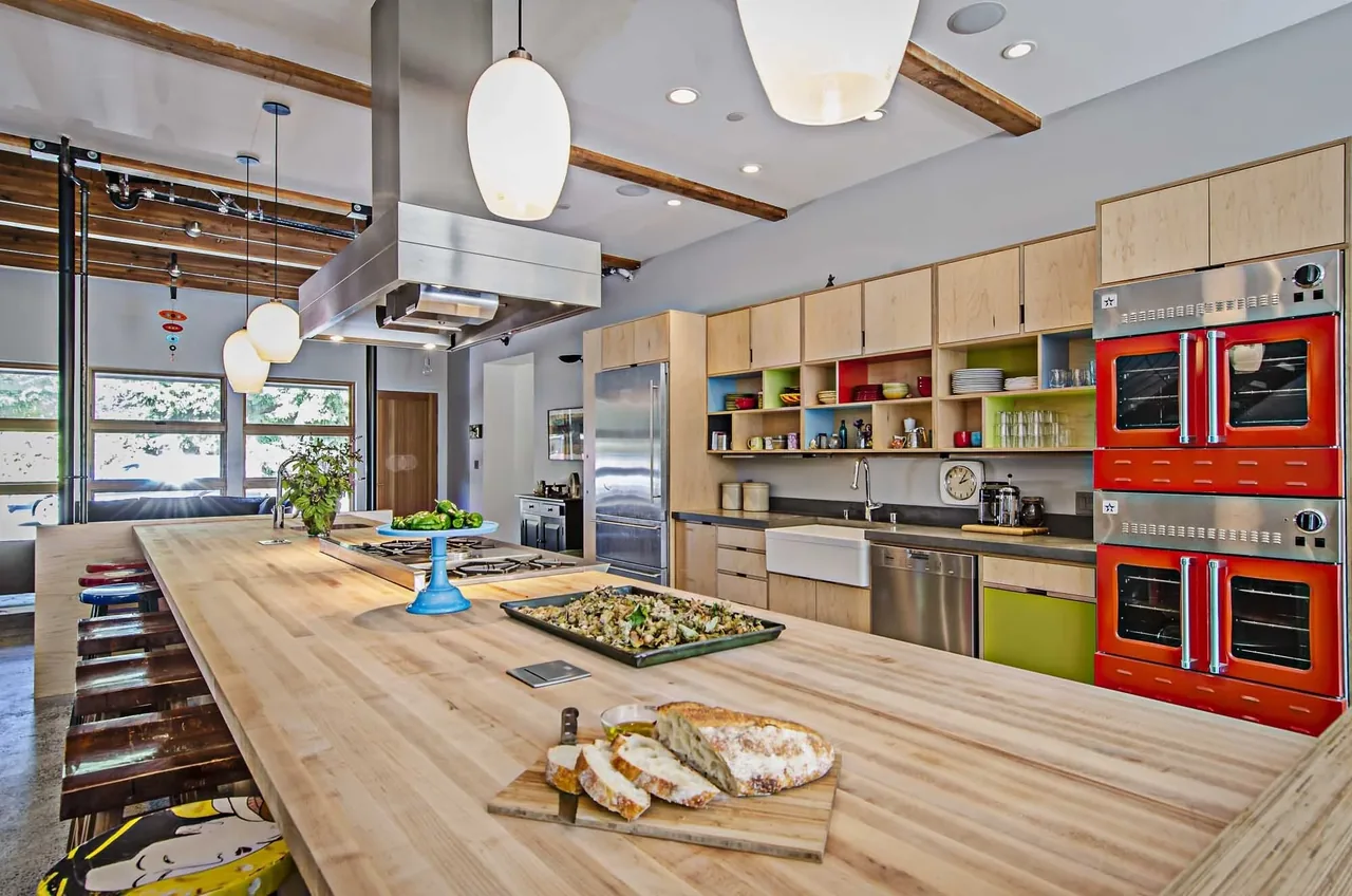 Kitchen interior with a large wooden island, red double ovens, and open shelving in birch cabinets.