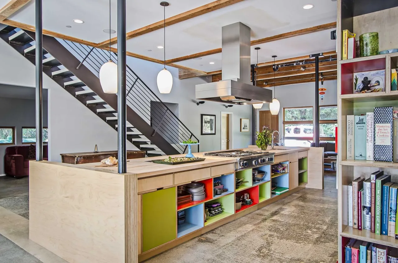 Modern kitchen with a wooden island featuring colorful cubbies, metal stair railing, and exposed ceiling beams.