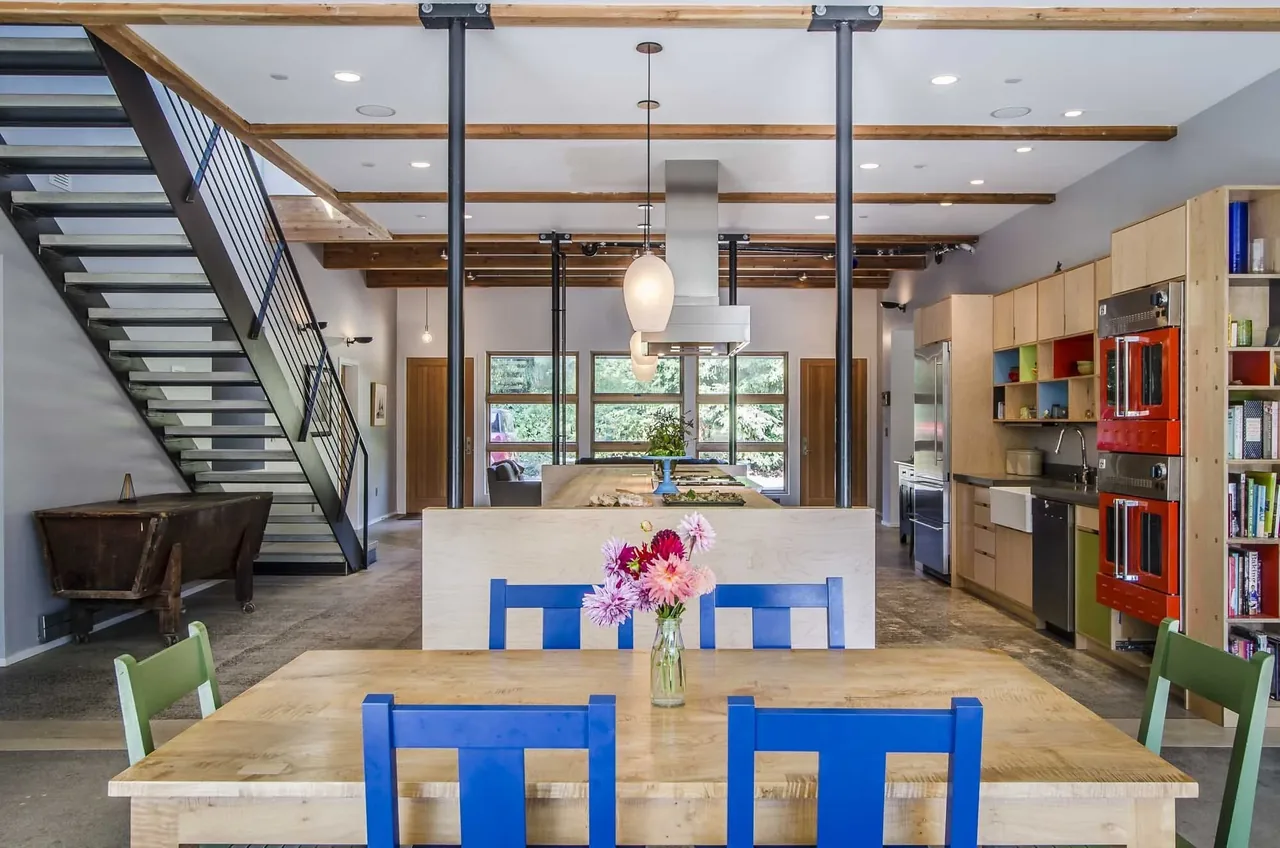 Open kitchen and dining space features exposed ceiling beams, metal stair railing, and red double ovens.