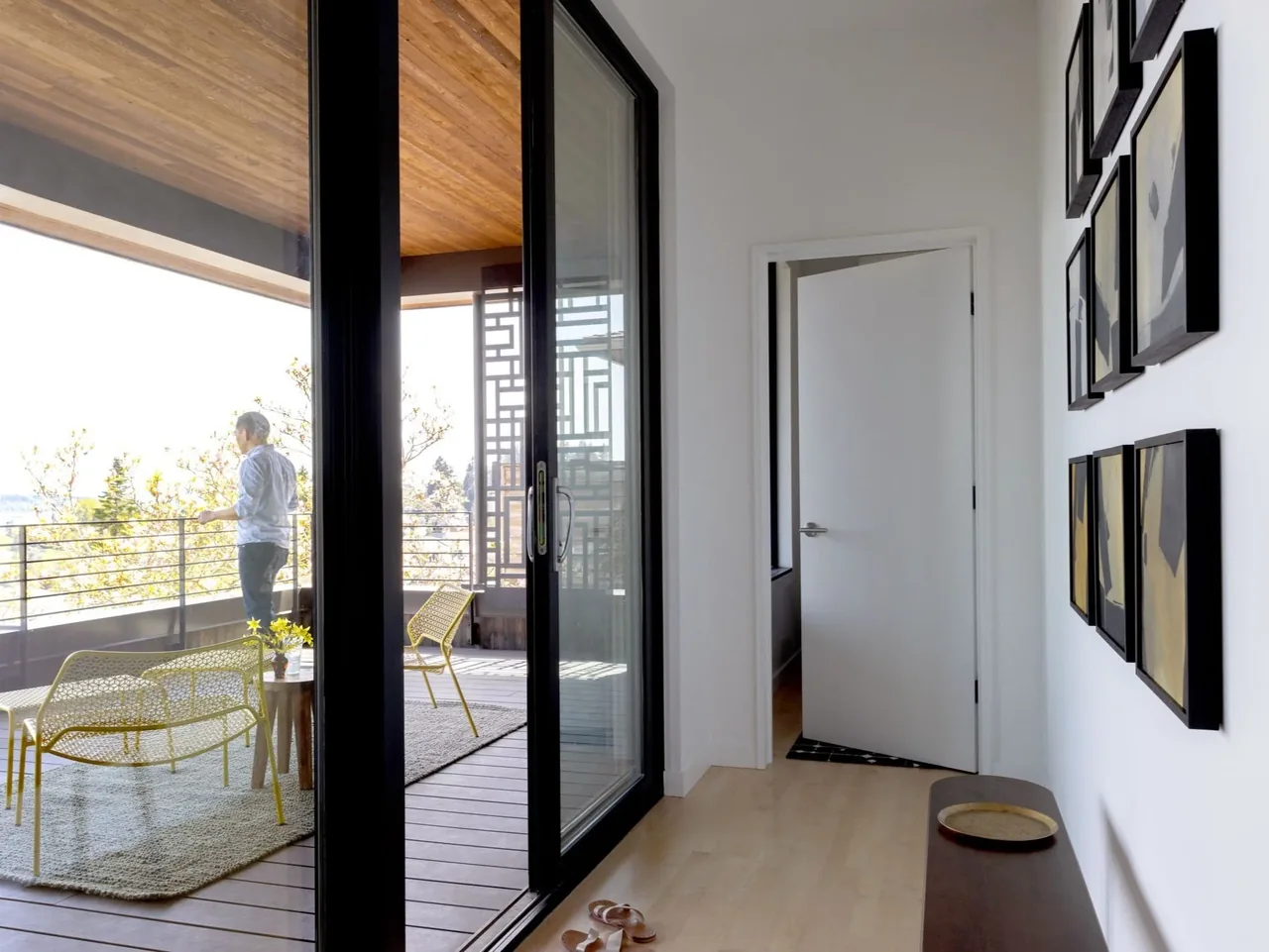 A hallway with light wood flooring and framed artwork leads to a covered patio with metal furniture and a scenic outdoor view.