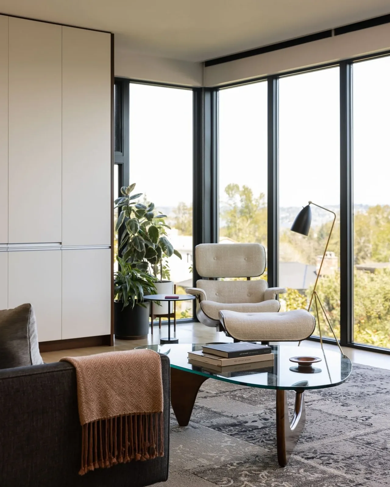 Bright living area showcasing a beige lounge chair and glass coffee table with books by large black-framed corner windows.