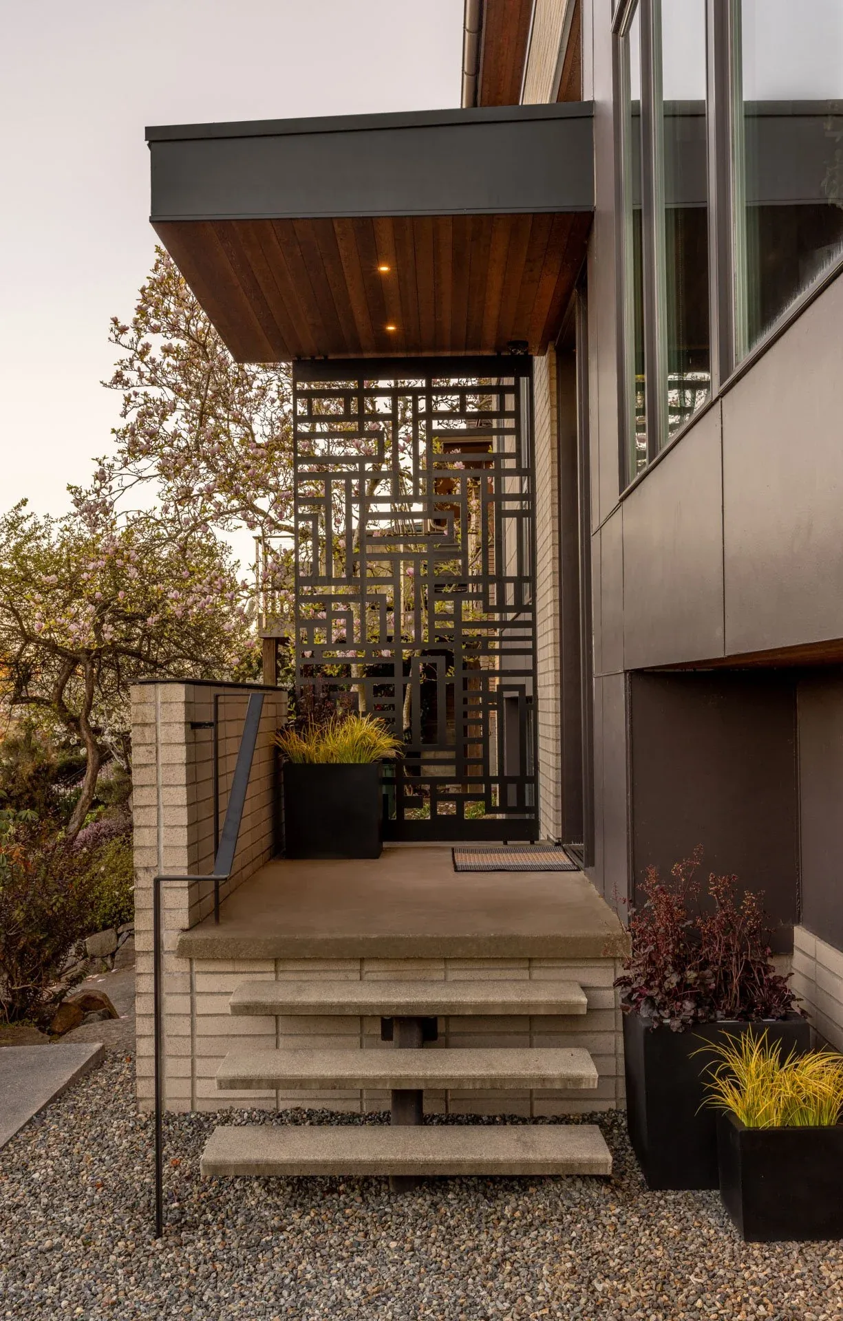 Modern home's entryway features concrete floating stairs, a wood-paneled ceiling overhang, and a geometric metal privacy screen.