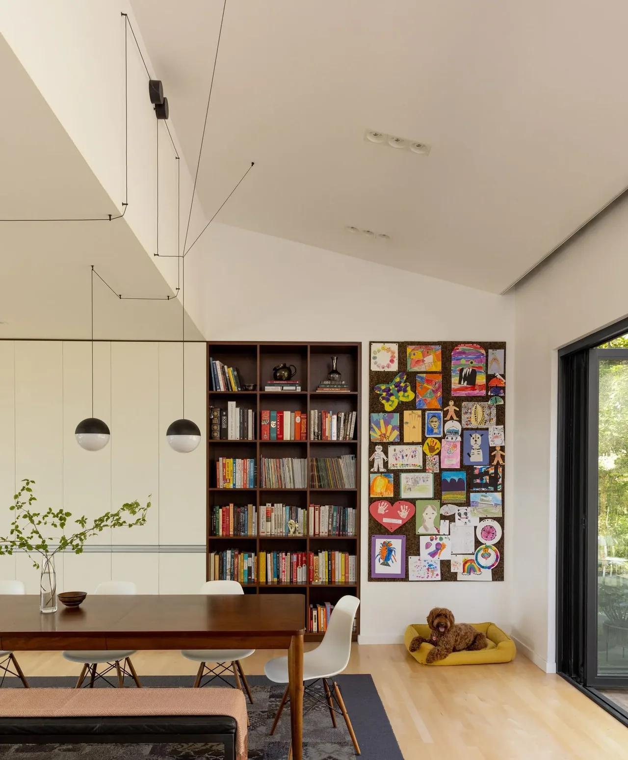 Dining area featuring a wooden table, white chairs, built-in bookshelf, and a wall displaying children's artwork in a light-filled space.
