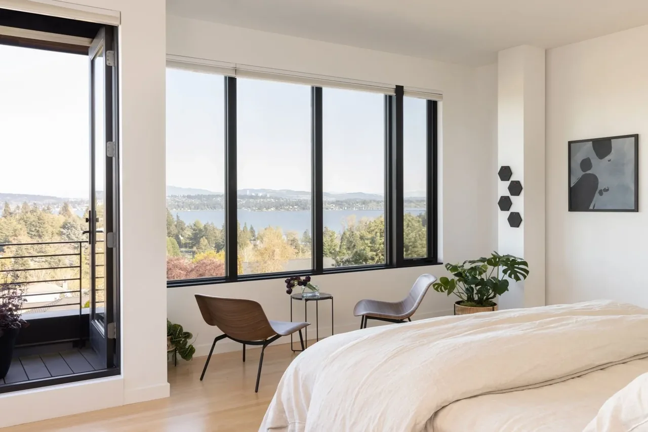 A bedroom featuring large black window frames with views of a lake and trees, plus a small seating area and balcony access.