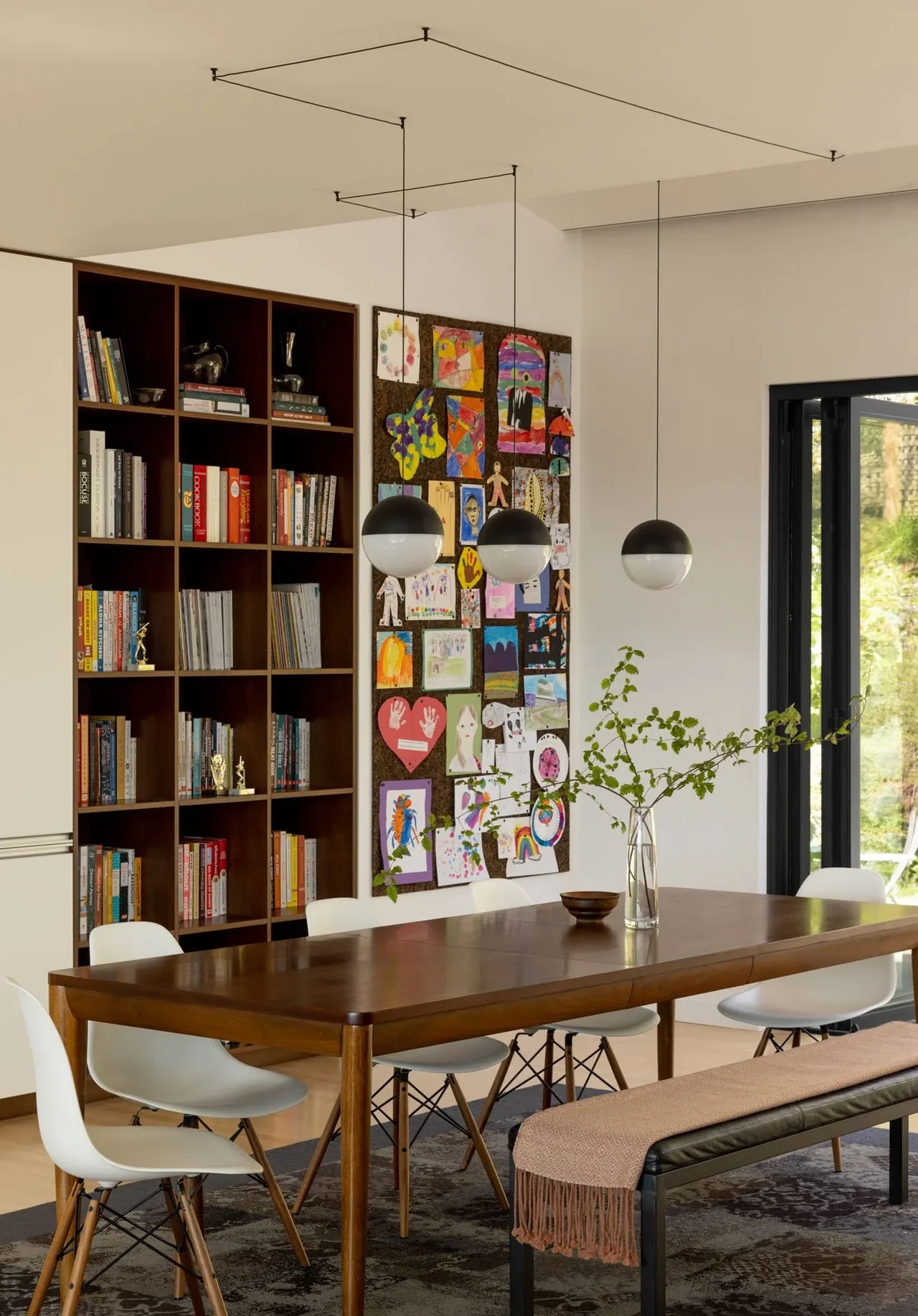 A dining space featuring a wooden table with white chairs and a black bench, pendant globe lights, and a large bookcase.