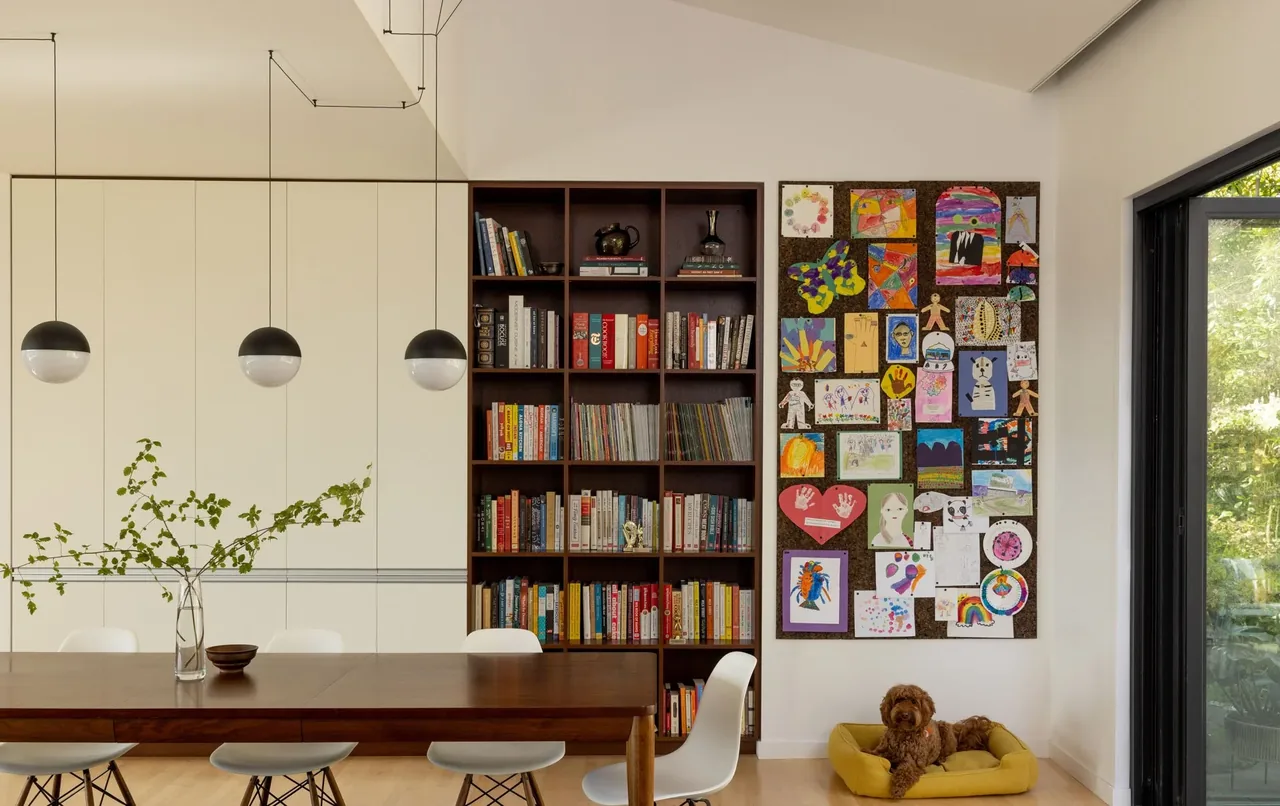 View of a dining area with pendant lights, dark wood table, built-in bookshelves, children's art display, and a dog bed by sliding glass doors.