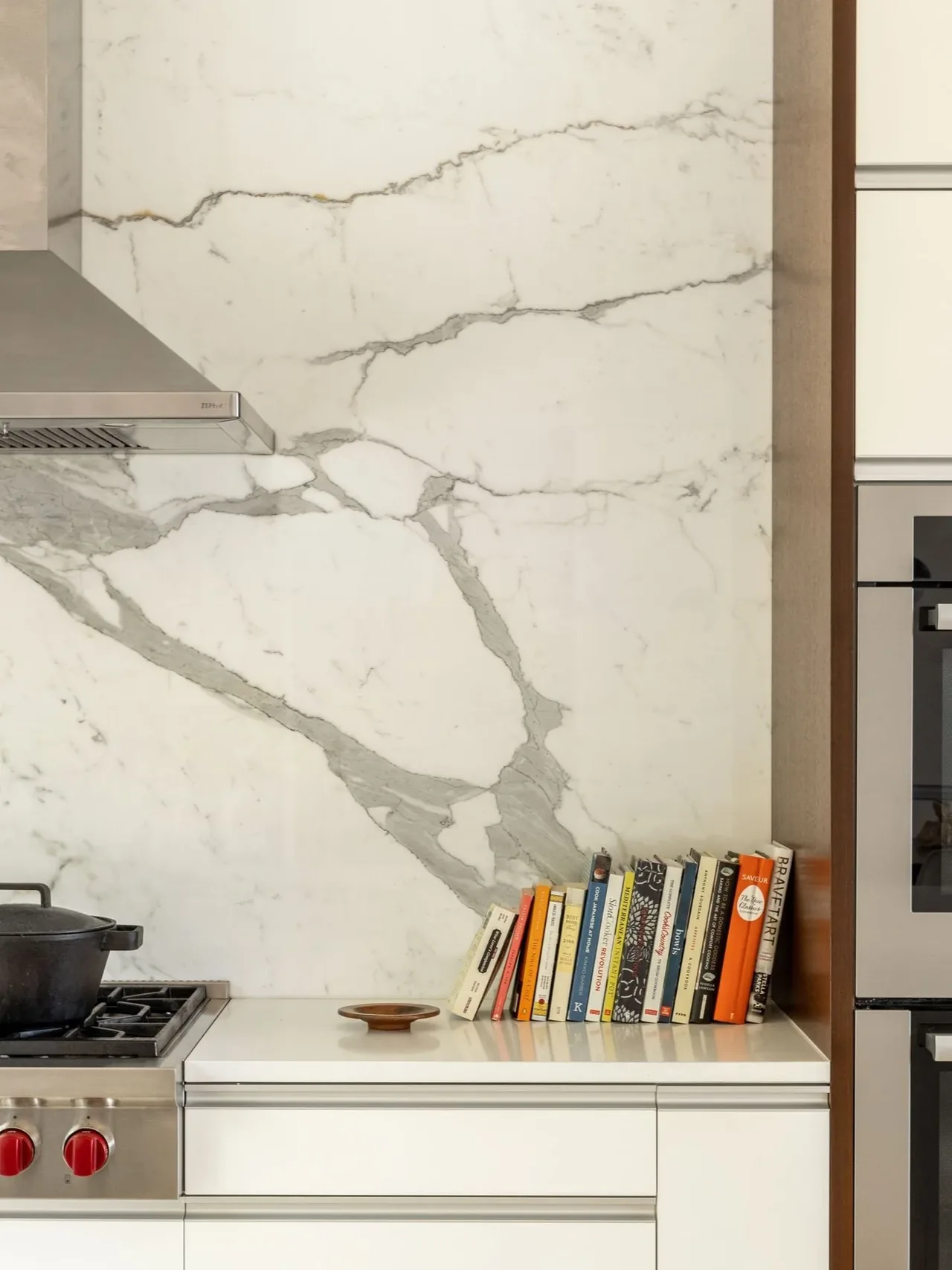 Marble backsplash with bold gray veining in a kitchen, accompanied by cookbooks on the white countertop.