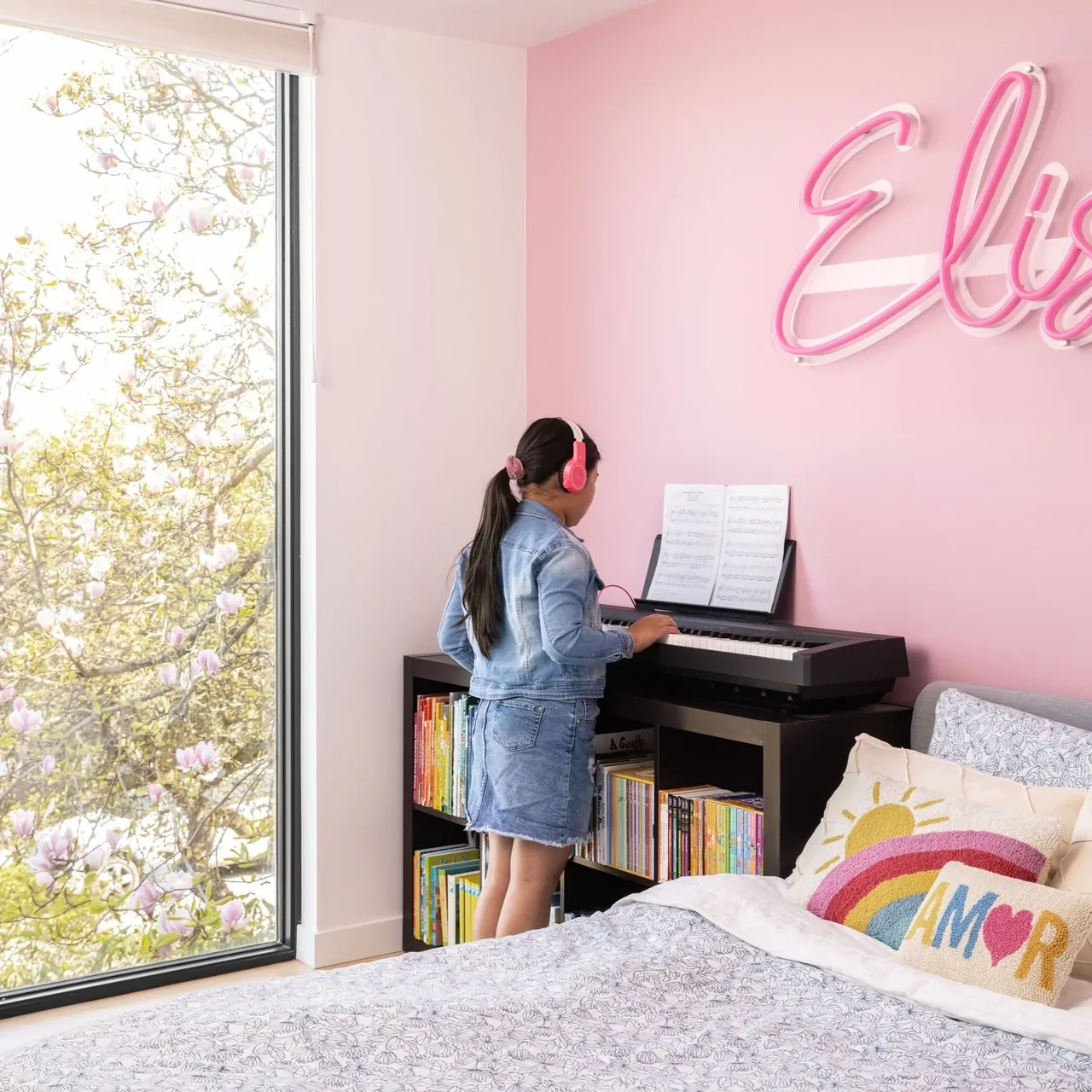 Child's bedroom features a keyboard, pink wall with script neon sign, and a large window with a view of blossoming trees.