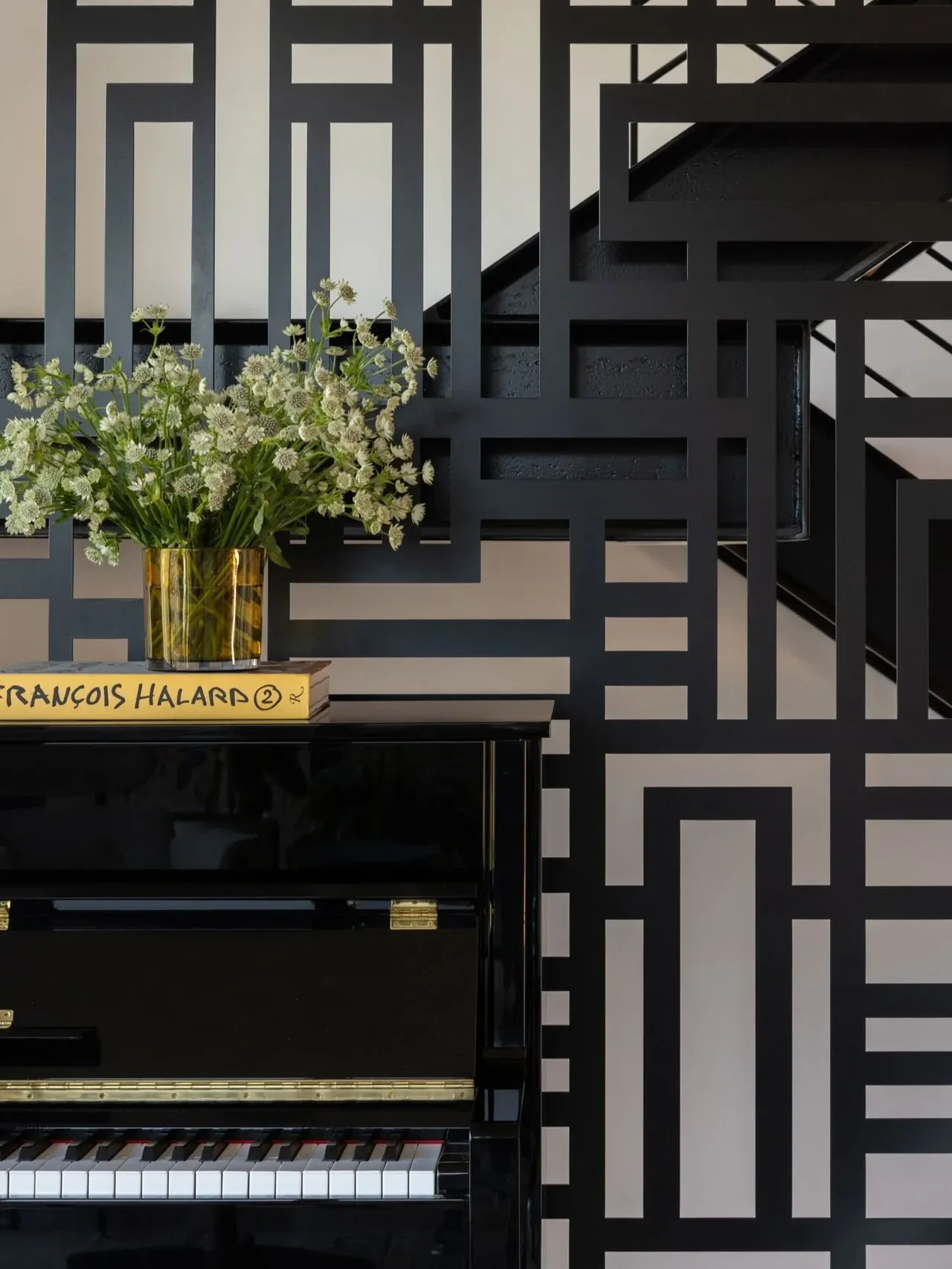 A black piano with a floral arrangement sits in front of a geometric patterned black wall feature near a staircase.