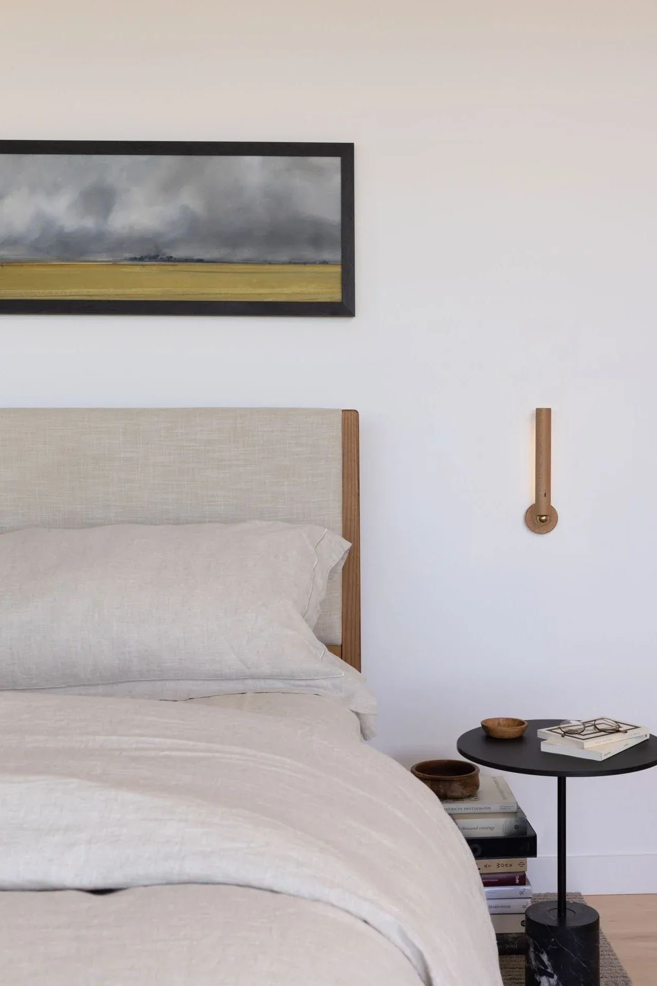 Minimalist bedroom featuring a light wood headboard, beige linens, a black side table, and a simple wooden wall sconce.
