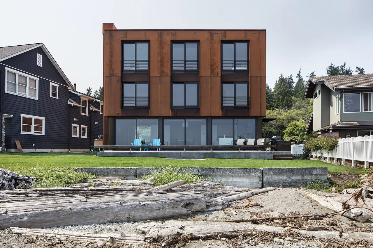 Whidbey Beach House features rust-colored metal exterior cladding and multiple large windows overlooking the waterfront.