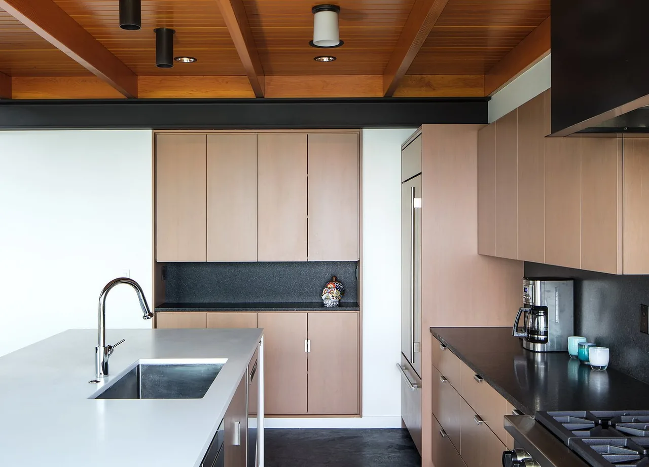 Modern kitchen featuring light wood cabinets, a white island with built-in sink, black countertops, and a wood-paneled ceiling with recessed lighting.