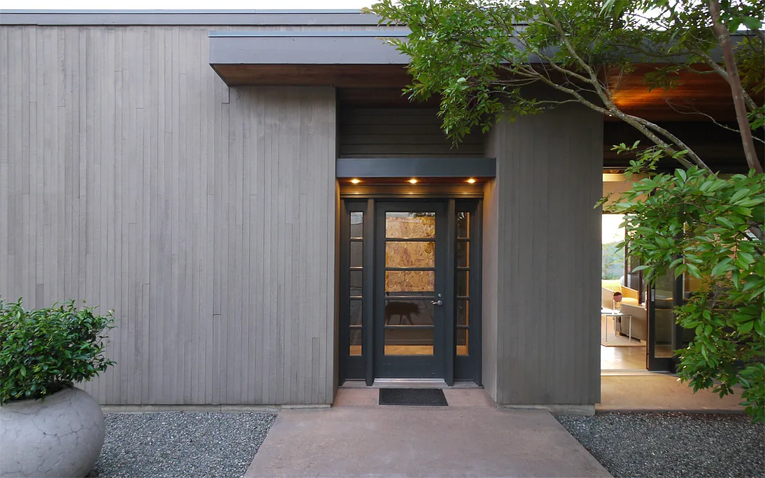 Front entry of a house featuring vertical wood siding and a black framed glass door under a flat roof with recessed lighting.