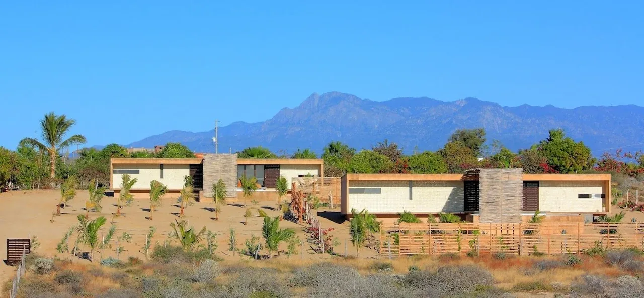 Modern flat-roofed homes with stone and wood elements are set on a desert-like landscape with mountain views.