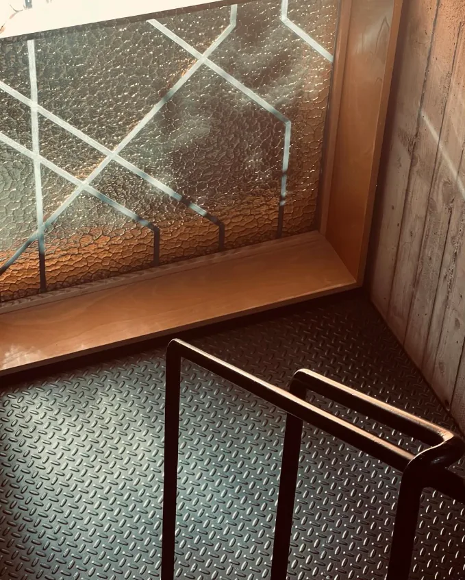 Textured glass window with wooden frame adjacent to black metal handrail and diamond-patterned rubber floor.