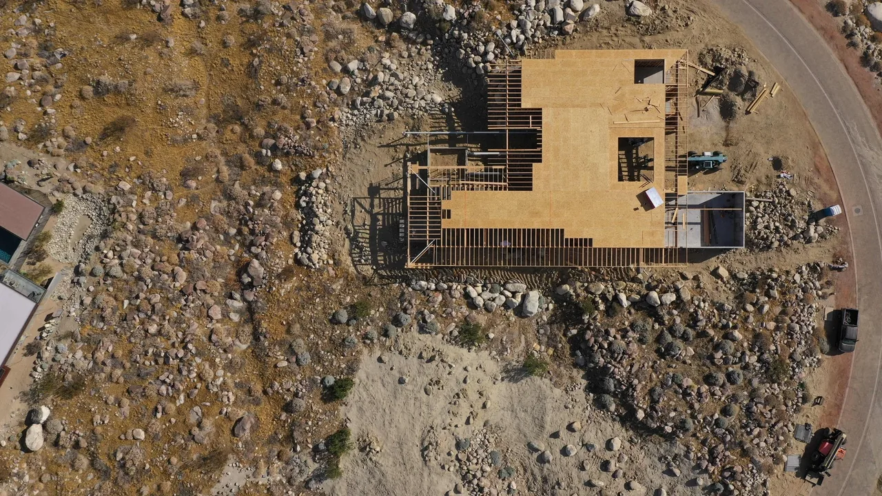 Overhead image shows the wooden framing of a house being built on a rocky lot near a curved street.