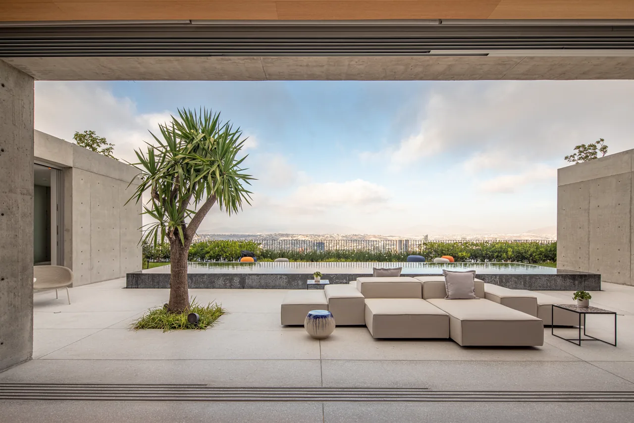 Concrete courtyard patio with a sectional sofa, small table, central tree, and pool overlooking the city.