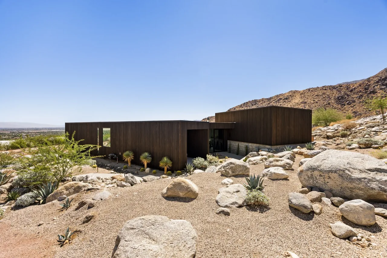 Modern house with a dark wood exterior sits on a rocky hillside with desert landscaping and large boulders.