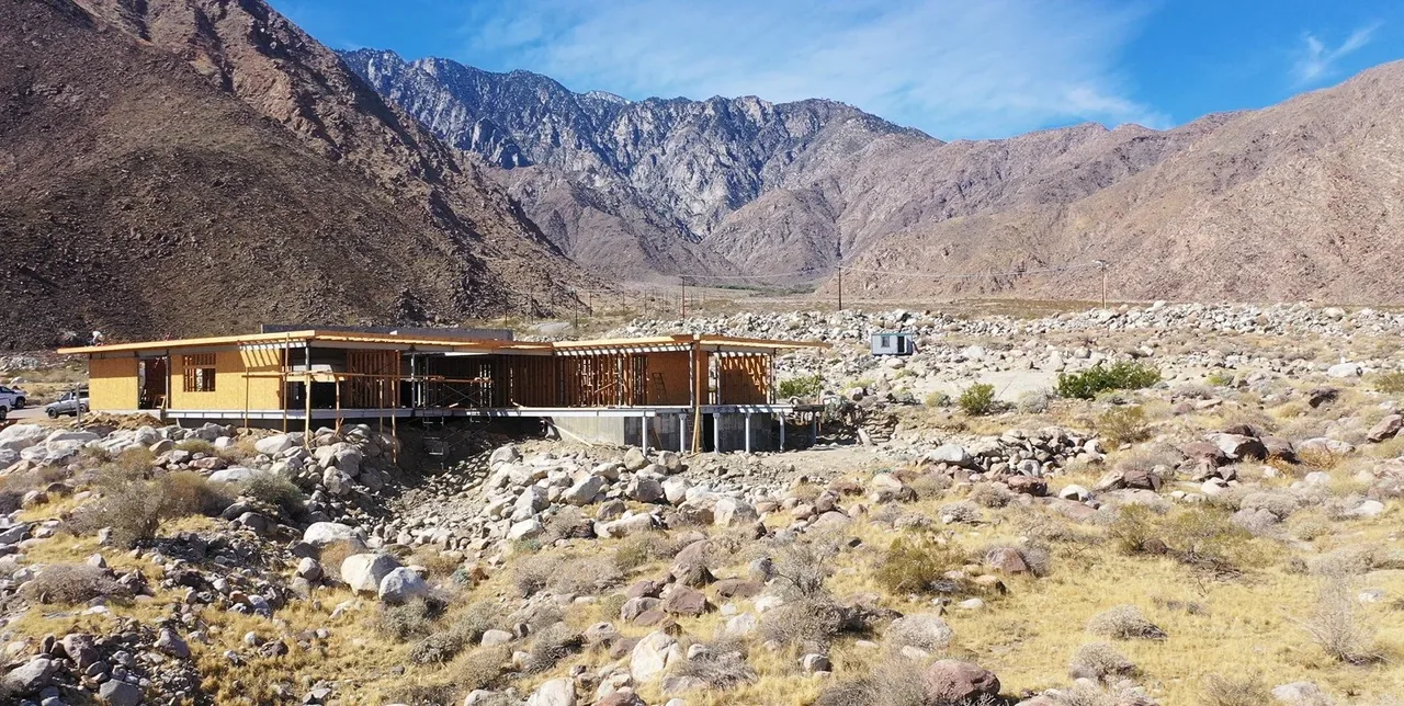 A modern home under construction in a rocky hillside desert landscape with mountain views.