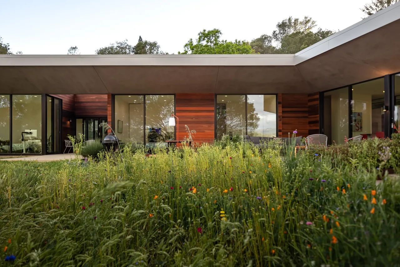Modern L-shaped home with floor-to-ceiling windows and rich wood siding opening to a lush wildflower garden.