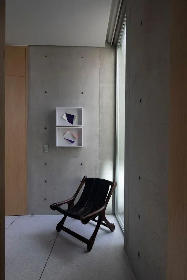 Minimal corner nook with a sling chair by a tall window and concrete walls featuring small geometric art.