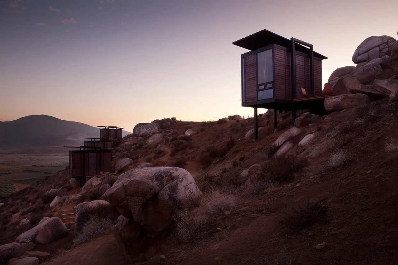 Modern cabins elevated on stilts set on a rocky hillside with a desert landscape and mountain backdrop at sunset.