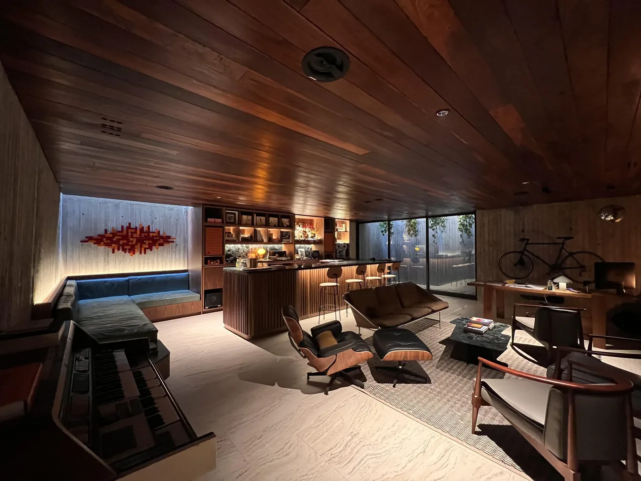 Interior living area with wood ceiling, built-in sectional sofa, bar counter with stools, and a piano in a modern design.