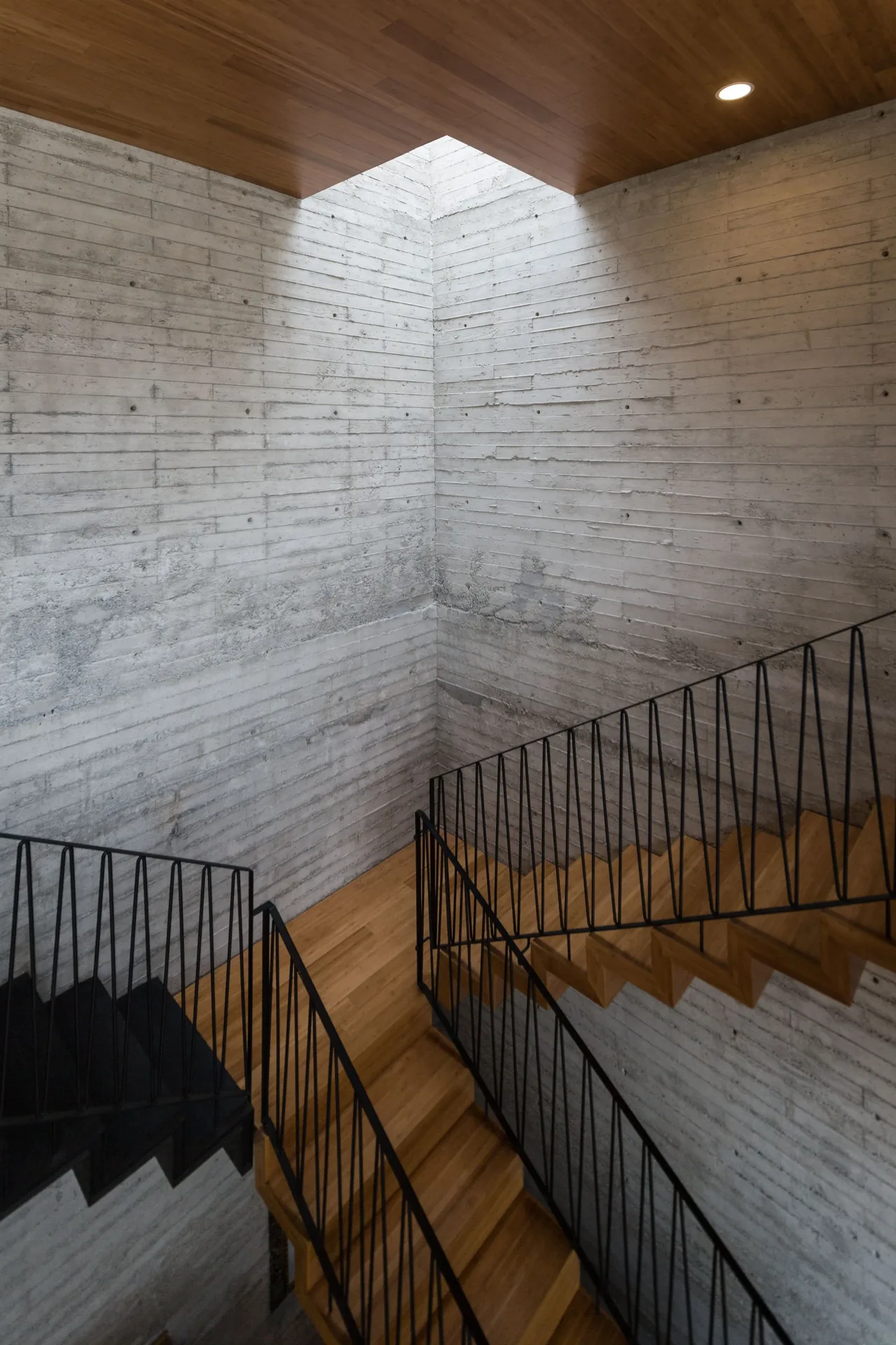 Stairwell featuring wood stairs, black metal railing, and board-formed concrete walls with a square skylight.