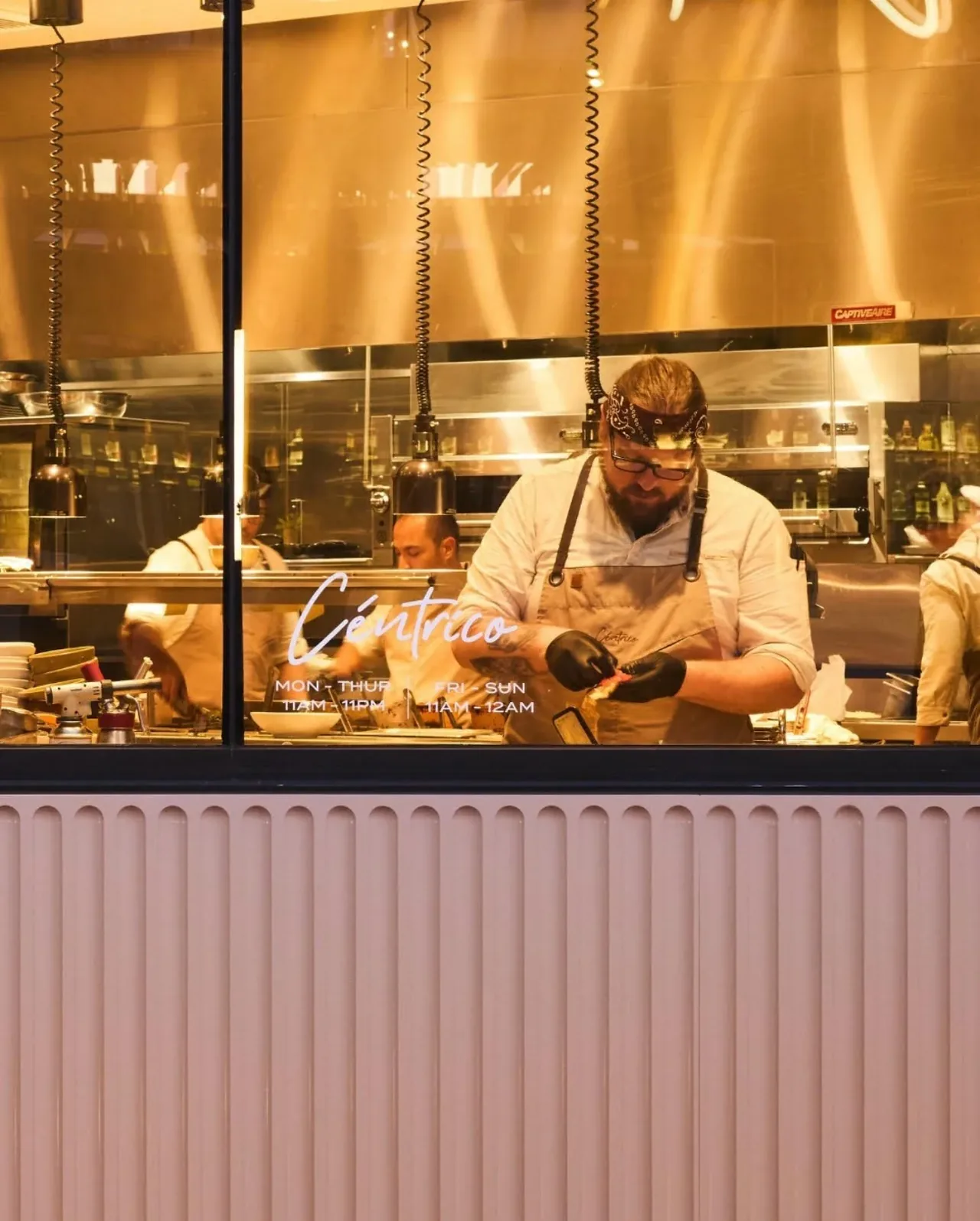 View through the front window into a commercial kitchen where a chef prepares food under hanging pendant lights.