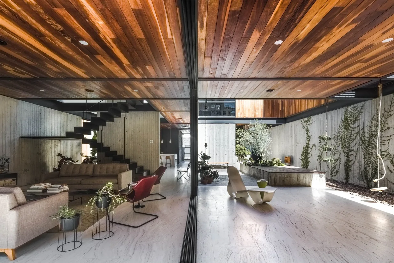 Living area with concrete walls, a floating staircase, glass partition, and wood-paneled ceiling illuminated by recessed lights.
