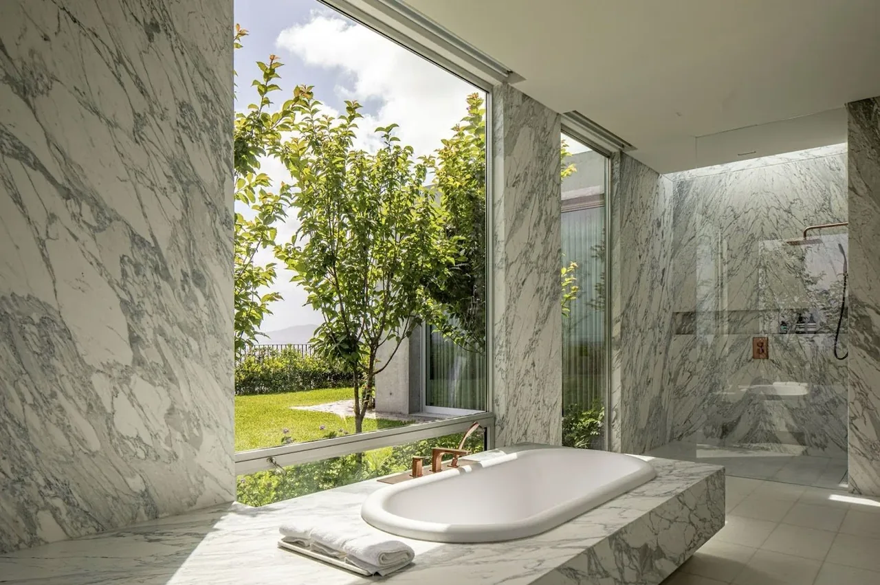 Bright bathroom with marble clad walls and a built-in bathtub facing large garden windows.