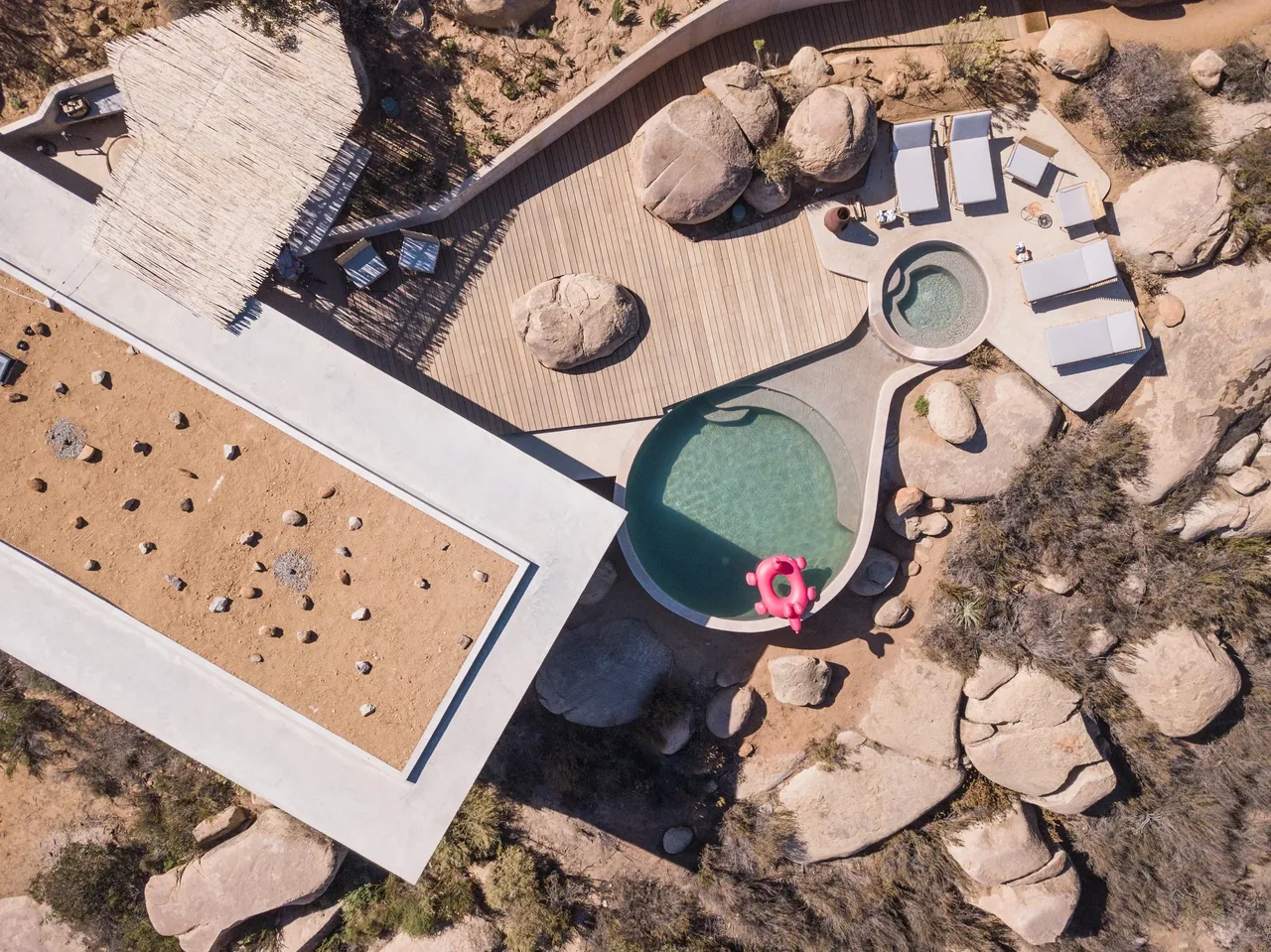 A kidney-shaped pool with a connected hot tub is integrated into a rocky desert landscape with a wooden deck and lounge seating.