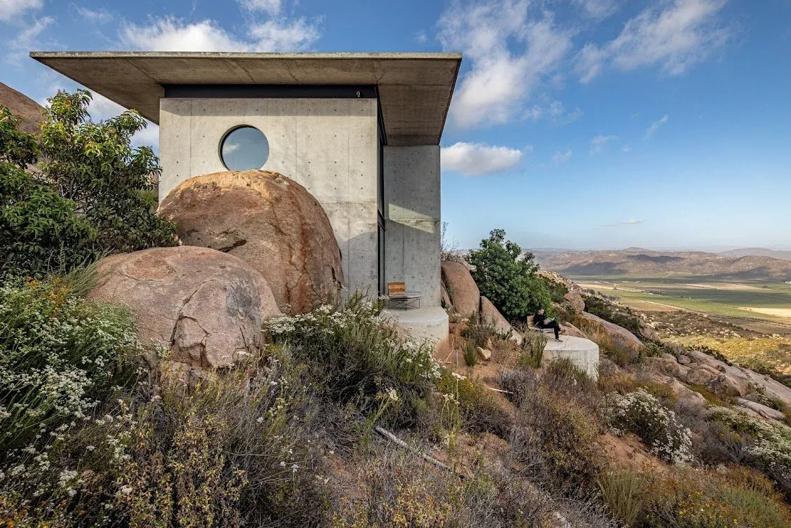 Concrete house by Gracia Studio integrates with boulders on a hillside overlooking the valley.