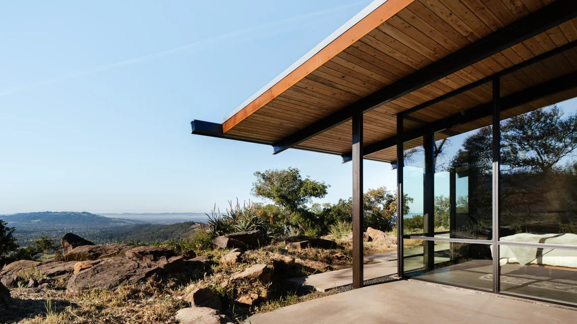 Modern home corner showing wood-paneled ceiling and expansive black-framed glazing overlooking a natural hillside.