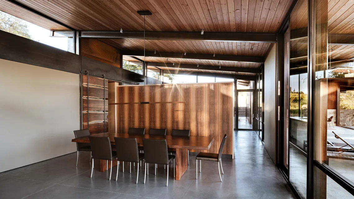 Dining room features wood-paneled ceiling and walls with a large wooden table and glass window views to outdoor spaces.