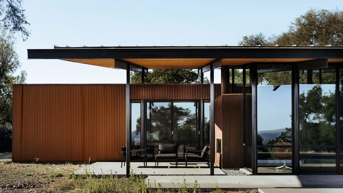 Modern house exterior featuring extensive black-framed glass walls and a wood-paneled ceiling on the patio cover.