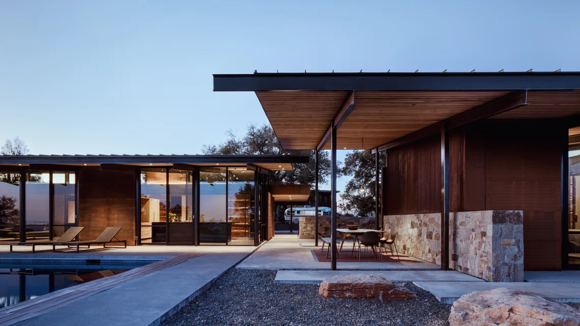 Contemporary house with floor-to-ceiling glazing and wood ceiling slats opening to a poolside patio with lounge chairs.