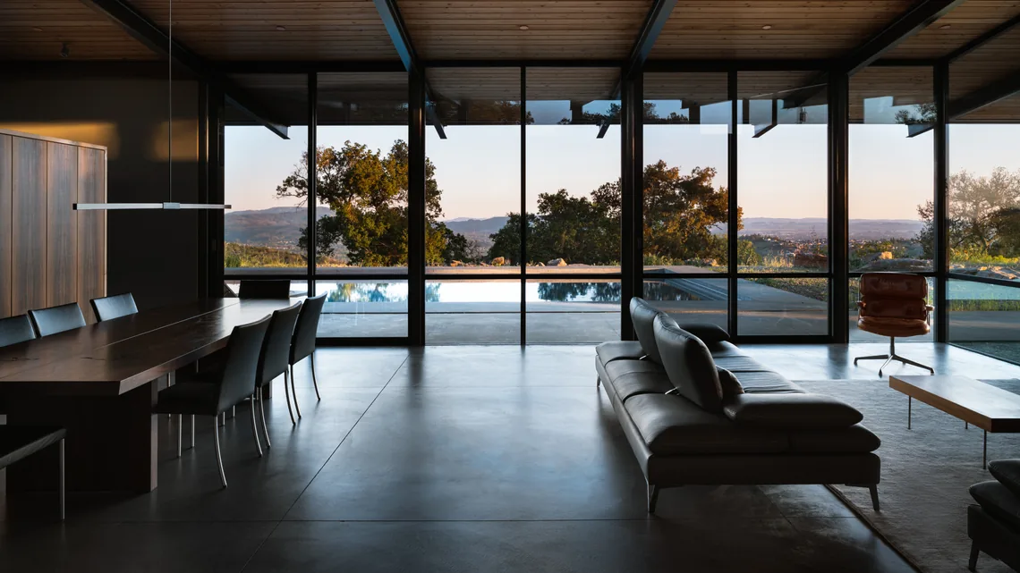 Modern living and dining space with glass walls framing a pool and nature outside at sunset.